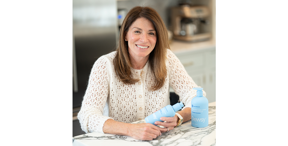 Dewsy founder Kathy smiling at a table with Dewsy Down Deep body care bottles — About Us founder portrait